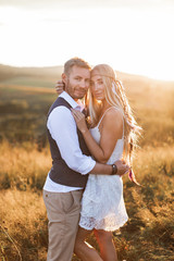 beautiful couple dressed in boho style embracing each other in the sunny summer field. Handsome man in casual wear and woman in white dress and feathers in hair walking in the field at sunset