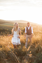 Stylish handsome man walking to his happy woman in boho clothes in sun light, romantic moment outdoors in the summer sunset field. Focus on the man