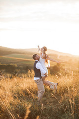 Young couple wearing hipster boho indie style clothes, in love walking in countryside, man holds woman on hands, woman is happy and smiling. Sunny sunset field on background, warm summer day