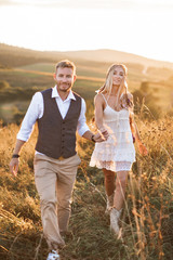 summer, love, romance and people concept - happy smiling young hippie boho couple holding hands and walking in summer field, outdoors. Woman in boho dress and hair feathers © sofiko14