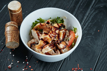 Appetizing salad with grilled chicken fillet, balsamic sauce, arugula and tomatoes in a white bowl on a black wooden background. Copy space, selective focus