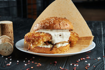 Appetizing and juicy burger with deep-fried fish fillet, white tartar sauce and cheese on a white plate with french fries.