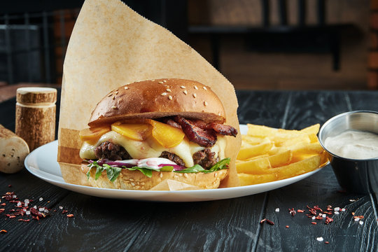 Appetizing and juicy burger with a large beef patty, melted cheddar cheese, arugula, onion, white sauce and peach on a white plate with french fries.