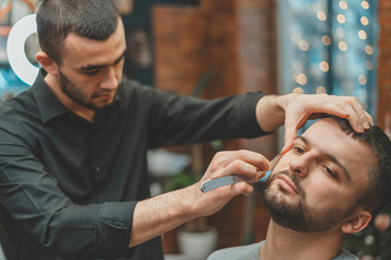 Obraz premium Beauty shop for men. Shaving a beard in a barbershop. Barber cuts his beard with a razor and clipper. close up Brutal haircuts. Hairdresser equipment. Selective focus.