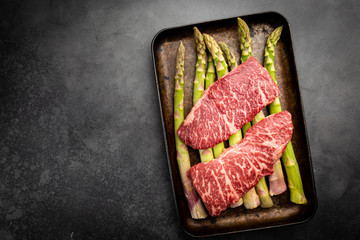 Raw Steak with green asparagus on tray on black background, top view
