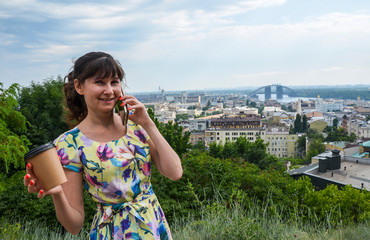 Fototapeta premium Beautiful young brunette woman in yellow dress talking on mobile phone and holding coffee and looking at the camera in fresh air