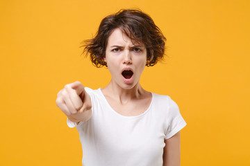 Angry young brunette woman girl in white t-shirt posing isolated on yellow orange background in studio. People lifestyle concept. Mock up copy space. Point index finger on camera swearing screaming.