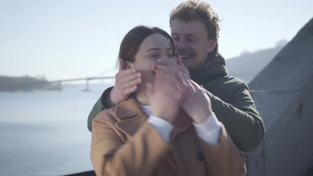 Young Caucasian man coming to girlfriend from background and closing her eyes. Excited chubby woman hugging boyfriend on river bank. Dating, love, romance, lifestyle.