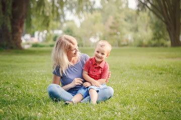Naklejka na ściany i meble Young smiling Caucasian mother and laughing boy toddler son sitting on grass in park. Family mom and child hugging having fun outdoor on a summer day. Happy authentic family childhood lifestyle.