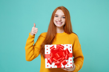 Smiling redhead girl in yellow sweater posing isolated on blue turquoise background. Valentine's Day Women's Day, birthday concept. Hold white red present box with gift ribbon bow, showing thumb up.