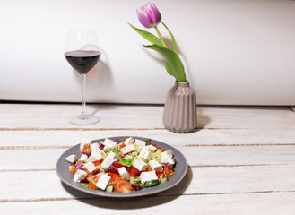 still life with plate of greek salad,  red wine glass, vase with tulip on white wooden table