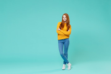 Smiling young redhead woman girl in yellow knitted sweater posing isolated on blue turquoise background studio portrait. People emotions lifestyle concept. Mock up copy space. Holding hands crossed.
