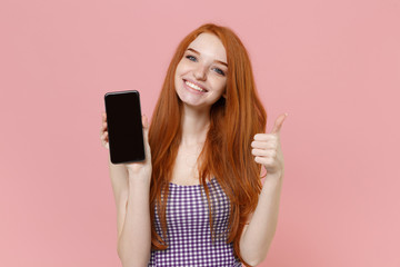 Smiling young redhead woman girl in plaid dress posing isolated on pastel pink background. People lifestyle concept. Mock up copy space. Hold mobile phone with blank empty screen showing thumb up.