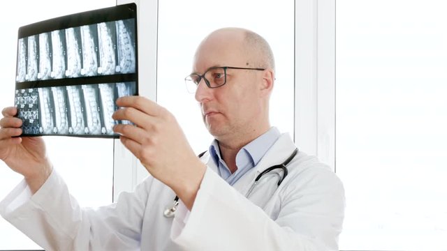 Man surgeon looking backbones x-ray front window in clinic office. Male doctor holding x-ray spine bones looking to camera. Portrait doctor in medical gown with phonendoscope