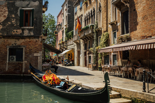 Charming landscape of typical street in Venice. Open-air cafe near canal with emerald water. Gondola parked near cafe. Sunny day.
