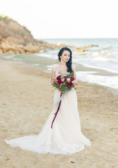 Fototapeta na wymiar Beautiful brunette bride in light chiffon wedding dress embroidered with beads posing near the sea. Romantic beautiful bride in luxury dress posing on the beach.