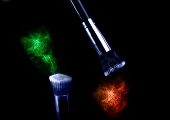 Two make up brushes with flying color powder, product on black background. Concept of glamour make up in minimal black.