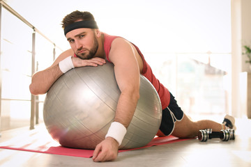 Lazy young man with exercise ball on yoga mat indoors