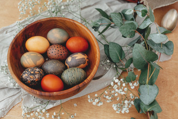Modern Easter eggs in wooden bowl with spring flowers and eucalyptus on rustic table. Stylish red, green and stone Easter eggs painted in natural dye. Happy Easter greetings.