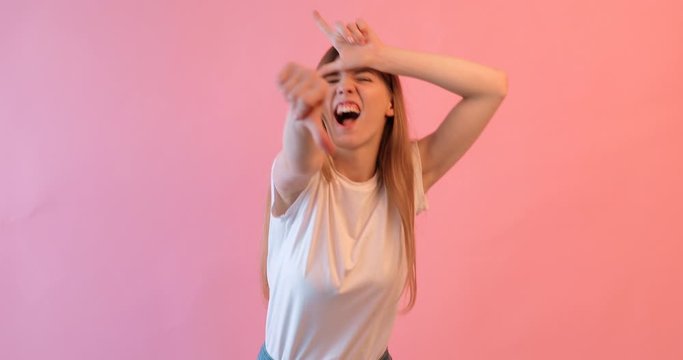 the girl shows the gesture of a loser or a loser , in a white t- shirt with a grin on her face, on a pink background