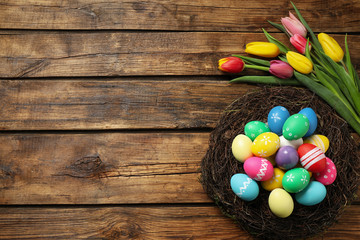 Colorful Easter eggs in decorative nest and tulips on wooden background, flat lay. Space for text
