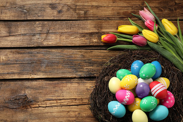 Colorful Easter eggs in decorative nest and tulips on wooden background, flat lay. Space for text