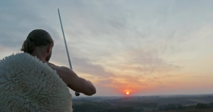 Man wearing a sheepskin holding a sword in his hand as he faces towards the sunset as the sun drops below the horizon raising the broadsword before making a slashing gesture in a rear view closeup -