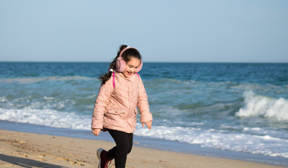 little girl in earmuffs and jacket run along the beach