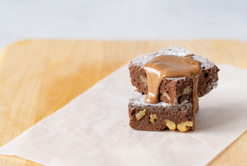 Two homemade brownie with nuts, icing sugar, melted chocolate on white layer of paper. Close up chocolate cake. Copy space. Wood background. Concept: pastry, American traditional dessert, bakery. 