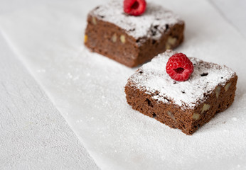 Two homemade chocolate brownie with nuts with icing sugar, strawberry on white paper. Close up chocolate cake. Copy space. Grey background. Concept: pastry, American traditional dessert, bakery.