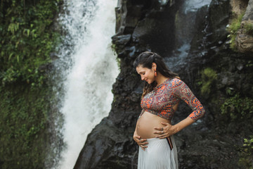 Portrait of young authentic pregnant woman near waterfall outdoors. Smiling and happy, holding belly. Spiritual harmony with nature. Travel and childbirth in Bali.