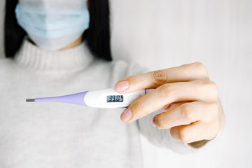 Young woman in medical disposable mask holds an electronic thermometer in her hands. High temperature on display