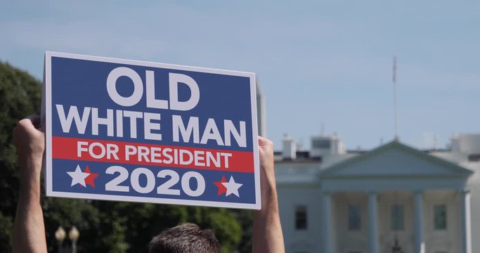 A man holds a fictional Old White Man for President 2020 election protest sign in front of the White House on a sunny summer day.	