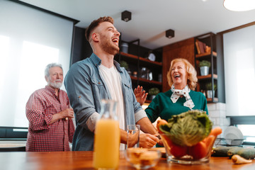 Young man is having good time while he preparing meal in the kitchen. He is laughing out loud while talking with his parents who are standing next to him.