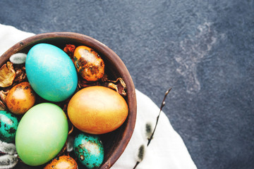 Bright easter eggs in a clay bowl and fluffy willow branches on a dark background, top view. Easter background. 	