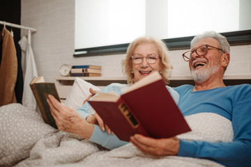 Portrait of good looking elderly couple wearing glasses are reading favorite books while lying in bed at bedroom.