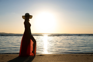 Young woman wearing long red dress and straw hat standing on sand beach at sea shore enjoying view...