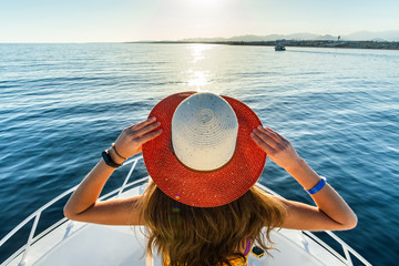 Young woman with long hair wearing yellow dress and straw hat standing on white yacht deck enjoying...