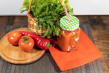 On the board are two tomatoes and red pepper. On a plate is a mug with tomato juice. In the background is a basket of green condiments..