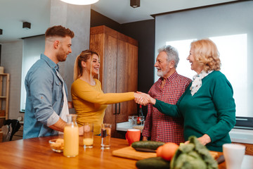 Obraz premium Four people are in the kitchen. Young blonde woman is shaking hands with elderly woman. Young and elderly man are standing next to them.
