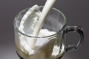 A stream of milk flows into a glass mug. Milk splashes at the bottom of the mug.