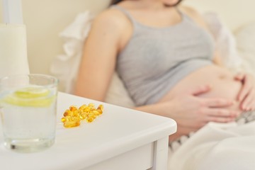 Obraz na płótnie Canvas Pregnant female in home interior sitting in bed, vitamin capsules close up