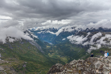 people stand on the edge of the mountain View to Geiranger fjord and eagle road in cloudy weather from Dalsnibba mountain, serpentine road, Norway, selective focus.