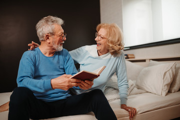 Portrait of elderly couple watching one another while sitting on bed in bedroom and reading book in the morning.