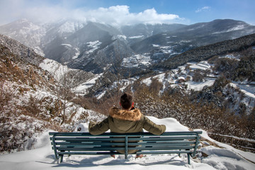 Young man sitting on a bench on the side of a mountain enjoying a snowy landscape in the background in winter. - obrazy, fototapety, plakaty