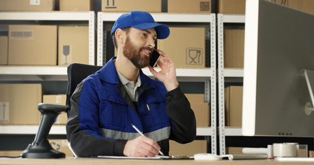 Caucasian man talking on mobile phone while working at post office store with parcels and on computer. Postman registering box and filling in invoice while speaking on cellphone in mail storage.