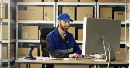 Handsome Caucasian mailman in blue iniform and cap sitting at table in postal office store and working at computer. Male post courier filling in invoice and entering data online in parcel storage.