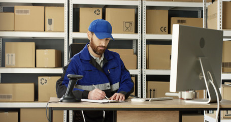 Handsome Caucasian mailman in blue iniform and cap sitting at table in postal office store and...