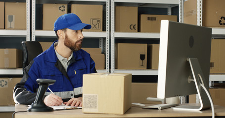 Handsome Caucasian mailman in blue iniform and cap sitting at table in postal office store and working at computer. Male post courier filling in invoice and entering data online in parcel storage. - obrazy, fototapety, plakaty