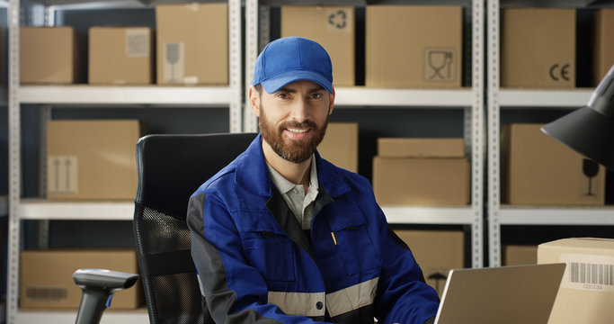 Portrait of young happy Caucasian postman in iniform and cap sitting at desk in postal office store and working at computer. Handsome male courier smiling happily to camera in storage of parcels.